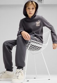 Sudadera gris con detalle de logo, pantalones de chándal gris a juego y zapatillas crema. Niño sentado en una silla de alambre blanca contra un fondo claro.