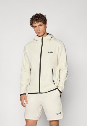 Bežna zip-up jakna s kapuco, črnimi detajli in dvema stranskima žepoma. V kombinaciji z ujemajočimi bež kratkimi hlačami z logotipom. Gladka tekstura.