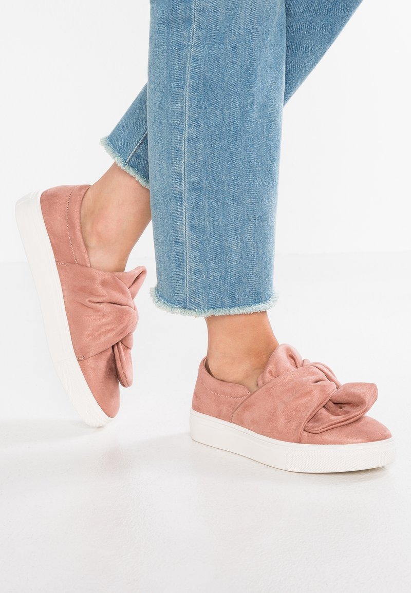 Personne portant un jean bleu clair à ourlet effiloché et des baskets roses à enfiler avec un grand nœud en tissu torsadé sur des semelles blanches.
