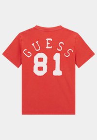 Sarkana kokvilnas T-kreklu ar baltu šenila izšuvumu, kurā virsū ir "GUESS" un apakšā "81", īsām piedurknēm un apaļu kakla dizains.