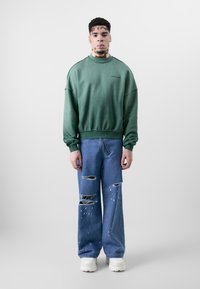 Grüner übergroßer Pullover mit gerippten Bündchen, kombiniert mit ausgewaschenen blauen Jeans mit großen Rissen und weißen Farbspritzern. Weiße Sneakers.
