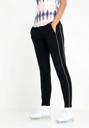 Person trägt schwarze Slim-Fit-Hose mit weißen Seitenstreifen, ein Batikshirt, weiße Sneakers, steht mit Händen in den Taschen.