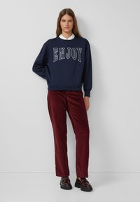 Marineblauwe sweatshirt met de tekst "ENJOY" in witte letters, gecombineerd met bordeauxrode corduroy broeken en glanzende donkere schoenen, met een casual pasvorm ontwerp.