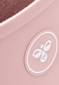 Conteneur rose rond avec un dessus brun texturé. Présente un logo circulaire blanc d'une abeille avec des gouttes et des feuilles. Surface en plastique lisse.