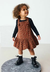 Braunes Corduroykleid mit schwarzem Leopardenmuster, gerafftem Saum und verstellbaren Trägern über einem schwarzen Langarmshirt. Schwarze Stiefel und weiße Socken.