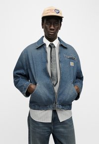 Νέος άνδρας που φοράει μπεζ καπέλο, υπερμεγέθη μπλε τζιν μπουφάν, άσπρο πουκάμισο, μαύρη γραβάτα και μπλε τζιν, στέ κεται με τα χέρια στις τσέπες.