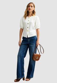 Witte blouse met knoopsluiting en pofmouwen, voorzien van gouden knopen. Gecombineerd met wijde blauwe denim broek en een bruine crossbody tas.