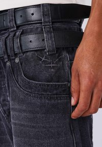 Vaqueros negros de mezclilla con un acabado texturizado, que presentan múltiples pasadores para cinturón, costuras visibles y un sutil bordado decorativo cerca de los bolsillos.