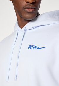 Biela mikina s kapucňou a sťahovacími šnúrkami, s modrým textom "INTER" a logom Nike na prednej strane. Hladký povrch a ležérny dizajn.