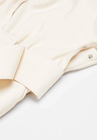 Blusa de un beige claro con una textura suave, que presenta un cuello ancho y un lazo decorativo en el costado, sujeto con un herraje de botón decorativo.