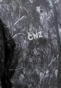 Nærbillede af en jakke med sort og grå mønster, med en lynlås foran og hvidt "CNZ" logo på brystet.