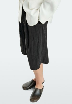 Personne portant un pantalon noir texturé à jambes larges et coupé, un blazer blanc, et des sabots en cuir noirs à enfiler avec une bride au talon.