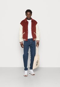 Giacca varsity bordeaux e crema con polsini a righe, t-shirt bianca, jeans blu e sneaker bianche. Borsa a tracolla beige con dettagli del logo neri.