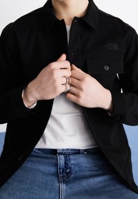 Veste noire boutonnée avec col et poches à l'avant. Portée sur un t-shirt blanc. Jean en denim bleu. Plusieurs bagues en argent aux mains.