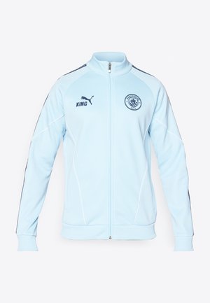 Chaqueta de chándal azul claro con cremallera y detalles en azul oscuro, con ribetes blancos y un logo bordado. Cuenta con puños y cinturilla de canalé.