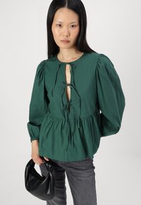 Blusa de algodão verde com mangas longas bufantes, decote redondo e detalhe de laço em forma de olho-chave na frente. Combinada com jeans escuros cinzentos e uma bolsa preta.
