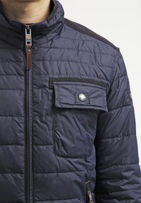 Primo piano di una persona che indossa una giacca trapuntata blu navy con una tasca sul petto a bottone automatico e una cerniera frontale.