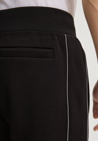 Czarne spodnie sportowe z teksturowanym materiałem, elastycznym pasem i dopasowanym krojem. Widoczna jest boczna kieszeń z detalem w postaci obszycia.