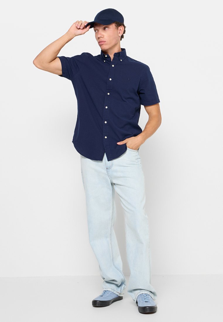 Chemise courte à manches avec boutons marins, jeans bleu clair et baskets bleues. La personne porte une casquette bleu marine assortie.