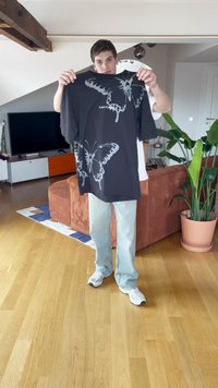 Svart t-skjorte med stort grått sommerfugl-trykk og teksten "TAK AV". Løs passform, korte ermer, glatt bomullstekstur.