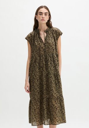 Femme aux longs cheveux foncés portant une robe fleurie ample sans manches avec un col froncé et une jupe à volants, debout devant un fond blanc.