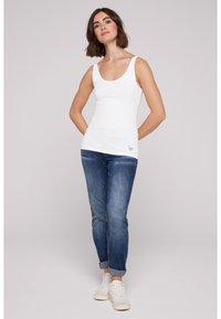 Top blanco sin mangas con escote redondo, combinado con jeans azules con los puños enrollados. El modelo lleva zapatillas blancas, mostrando un atuendo casual.
