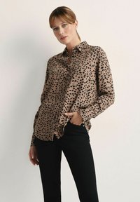 Brązowa bluzka w łaty z kołnierzykiem, zapinana na guziki z przodu, z długimi rękawami i luźnym krojem, sparowana z czarnymi spodniami. Gładka tekstura.