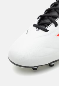 Botas de fútbol blancas con una superficie texturizada, cordones negros y tacos de goma negros. Presentan acentos rojos y una marca cerca del dedo del pie.
