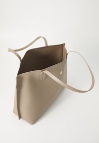 Taupe neplederen totebag met twee lange handvatten, open bovenkant en gouden logo-detail op de voorkant. Glad oppervlak met een minimalistisch ontwerp.