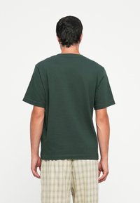 Tricou din bumbac de culoare verde închis, cu mâneci scurte și o croială lejeră, având un gât rotund clasic și un tiv drept, purtat împreună cu pantaloni în carouri.