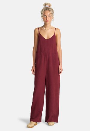 Bordowy kombinezon z pogniecionej tkaniny z cienkimi ramiączkami, dekoltem w serek, dwiema przednimi kieszeniami i szerokimi nogawkami, w połączeniu z drewnianymi butami na platformie.