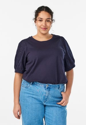 Mujer con cabello rizado que lleva una camiseta azul marino de manga corta con mangas abullonadas y vaqueros de tiro alto en azul claro, de pie frente a un fondo blanco.
