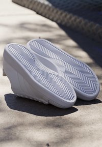 Par hvide kile-sneakers med detaljeret sildeben-mønster i sålerne, der hviler på betonoverflade i sollys.