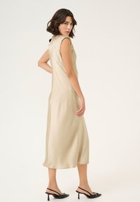 Vestito beige senza maniche con corpino aderente e gonna fluida; realizzato in tessuto morbido. Abbinato a sandali neri con tacco.