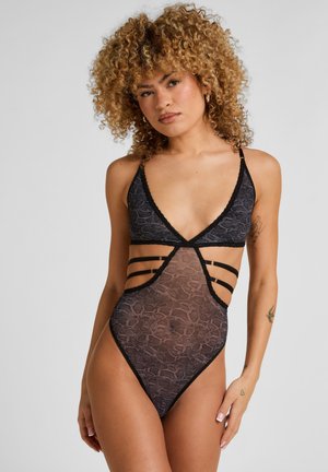 Vrouw met krullend haar, gekleed in een zwart kanten body met openingen en bandjes aan de zijkanten, staand tegen een effen lichtgrijze achtergrond.