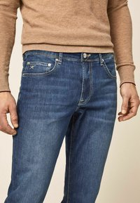 Hombre vestido con jeans de mezclilla azul y un suéter de manga larga beige, con las manos relajadas a los lados, frente a un fondo neutro.