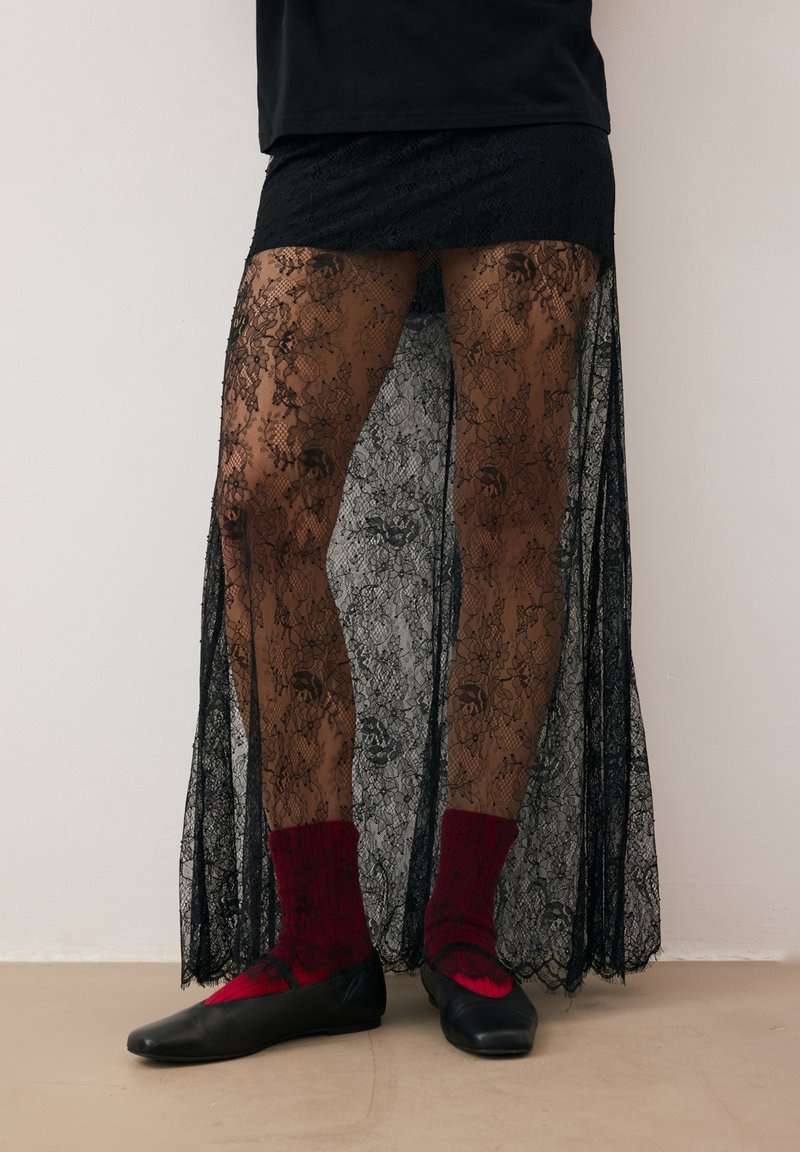 Czarna koronkowa spódnica z kwiatowym wzorem, półprzezroczysty materiał i boczne rozcięcia, zestawiona z czerwonymi skarpetkami i czarnymi balerinami. Prosty, elegancki design.