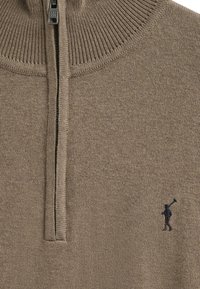 Brun stickad tröja med halvvide dragkedja och ribbad krage. Har en liten mörk logotyp av en figur som håller i en pinne. Mjuk textur, avslappnad design.