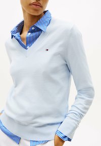 Pullover azzurro chiaro con scollo a V, realizzato in un tessuto morbido. Presenta polsini e orlo a costine. Sotto, un colletto a righe blu e bianche.
