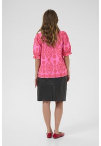 Blusa rosa con motivo a pallini e maniche a sbuffo abbinata a una gonna di pelle nera. Ballerine rosse completano l'outfit, evidenziando forti contrasti di colore.