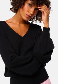 Pull noir en tricot avec un col en V, des manches ballon et des poignets côtelés ; texture douce et coupe décontractée. Le mannequin pose avec les bras croisés.