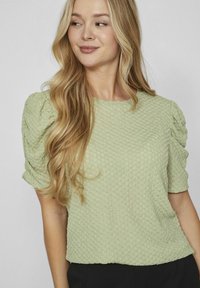 Lichtgroene, gemêleerde top met korte, opbollende mouwen, versierd met een honingraatpatroon en een ronde halslijn, gecombineerd met donkere broek.