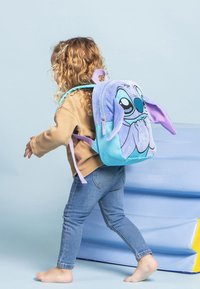 Sac à dos en peluche bleu et violet avec un design de personnage, de grandes oreilles et un détail de fermeture éclair. Porté par un enfant en pull marron et jeans bleus.