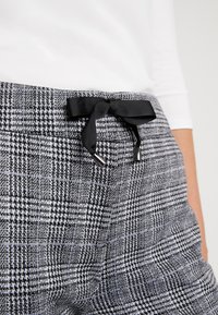 Spodnie w czarno-białą kratę o gładkiej fakturze, z czarną wstążką w talii i dwoma małymi srebrnymi akcentami.