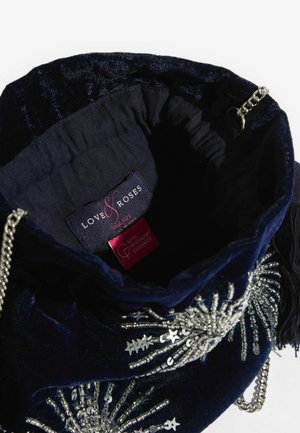 Tumesinine sametist tõmblukk-kotike, millel on hõbedast helmeste kaunistus ja kettpandlaga rihm. Sisemisel voodil on brändi silt.