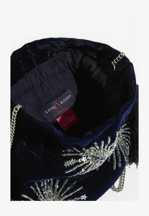 Tumesinine sametist tõmblukk-kotike, millel on hõbedast helmeste kaunistus ja kettpandlaga rihm. Sisemisel voodil on brändi silt.