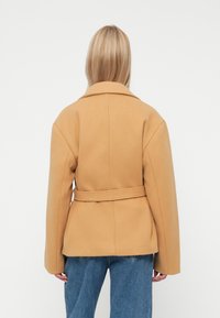 Karmelowy wełniany płaszcz z paskiem, okrągłym kołnierzem, długimi rękawami i wyraźnie zarysowaną sylwetką. Miękka tekstura; minimalistyczny design.