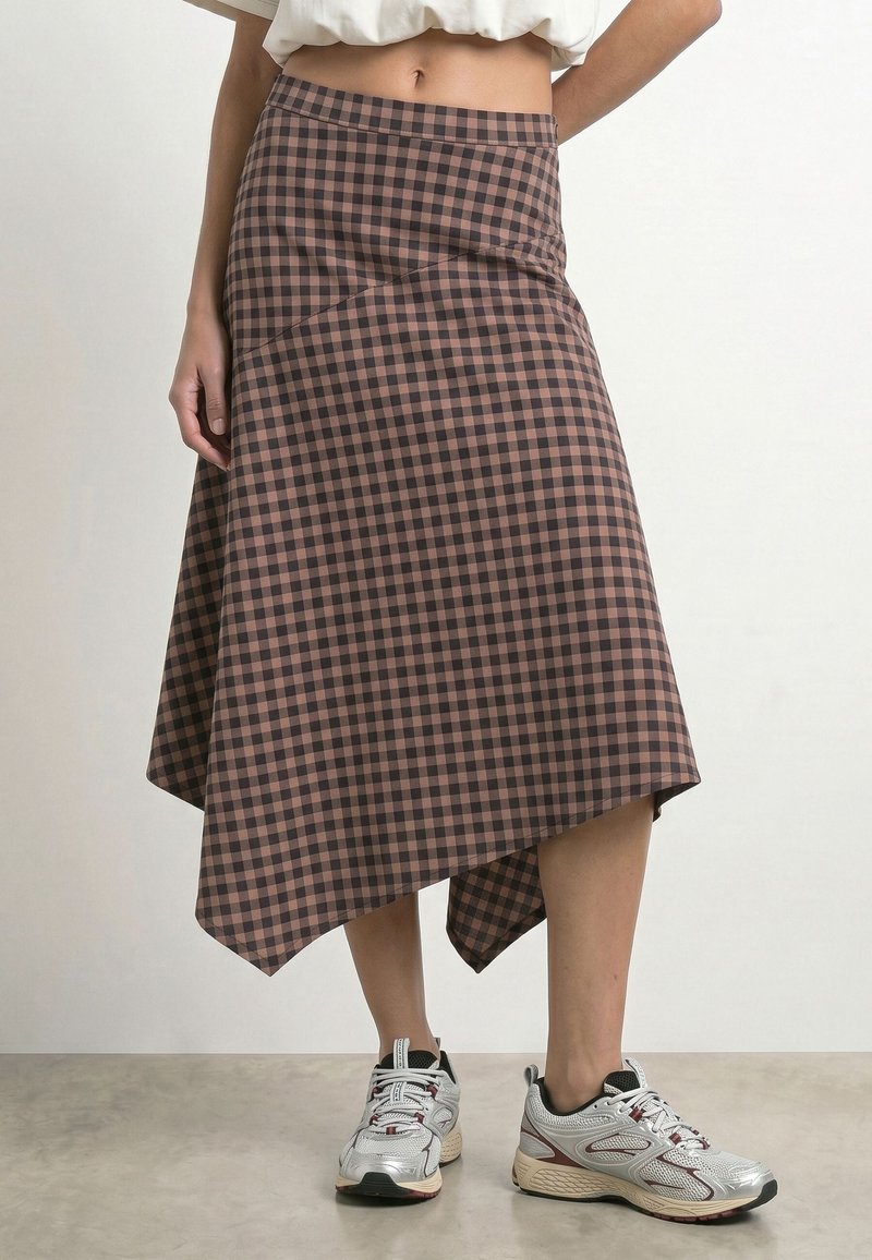 Personne portant une jupe asymétrique à carreaux marron et noir taille haute avec des baskets blanches et grises, debout devant un fond uni.