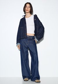 Jeans jakna s kratkim krojem in zapiranjem na gumbe, v kombinaciji s širokimi jeans hlačami. Oba kosa sta temno modra z kontrastnimi šivi.