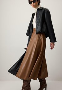 Črna usnjena kratka jakna, kremasti puli, rjava nagubana usnjena krilo in rjavi škornji do kolen na nevtralnem ozadju.